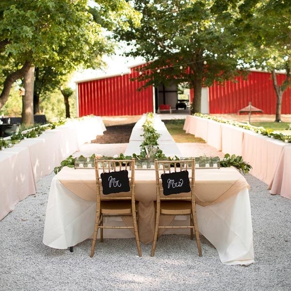 Elegant outdoor wedding reception setup with long tables, greenery, and rustic chairs.