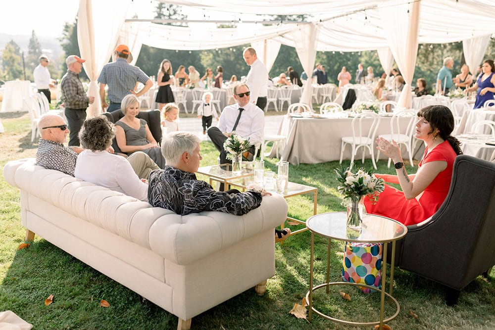 Elegant outdoor bridal event with guests socializing under a white tent, lush garden setting.