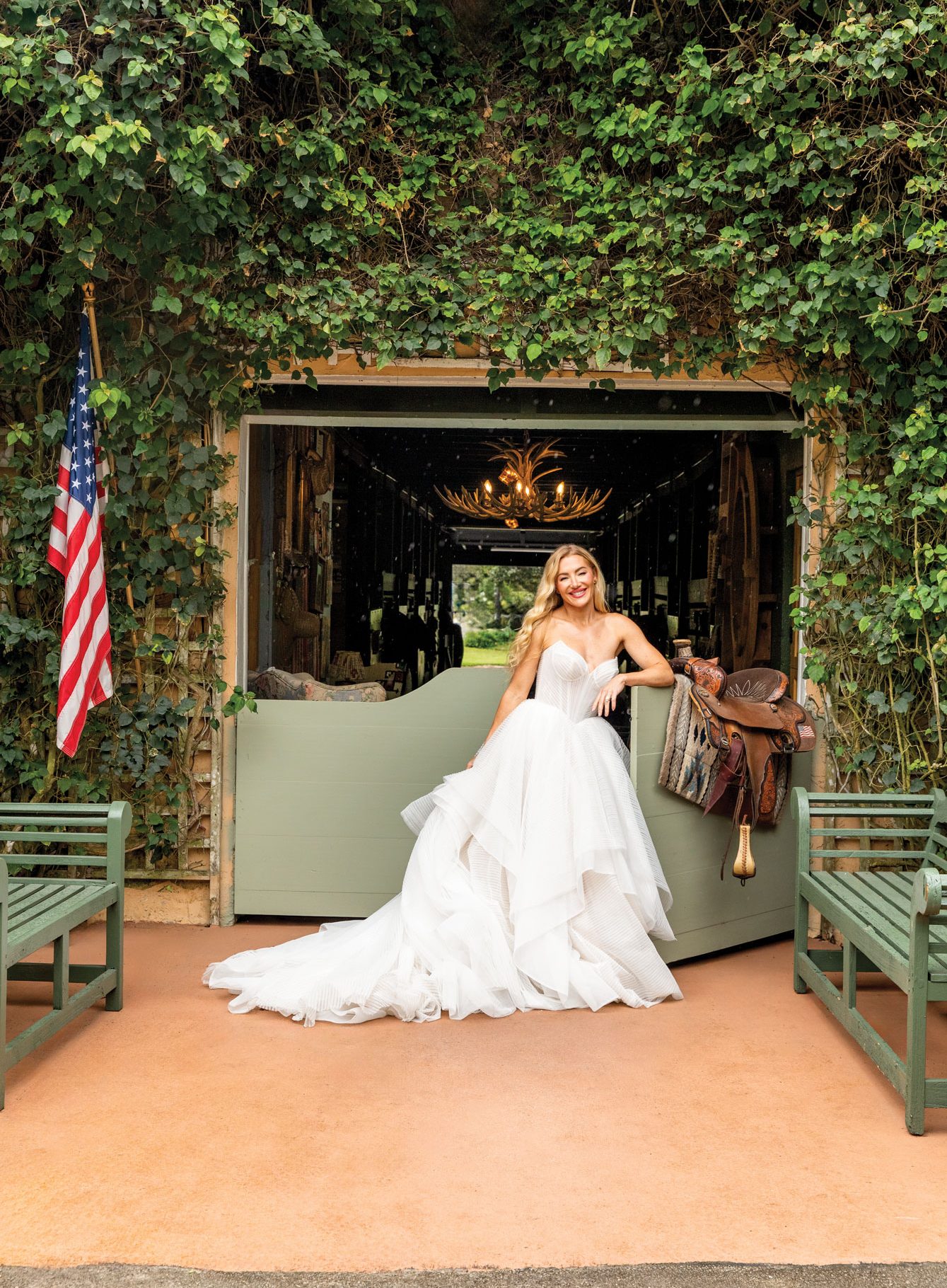 Elegant bride in a white wedding gown standing at the entrance of a rustic venue with greenery and A.