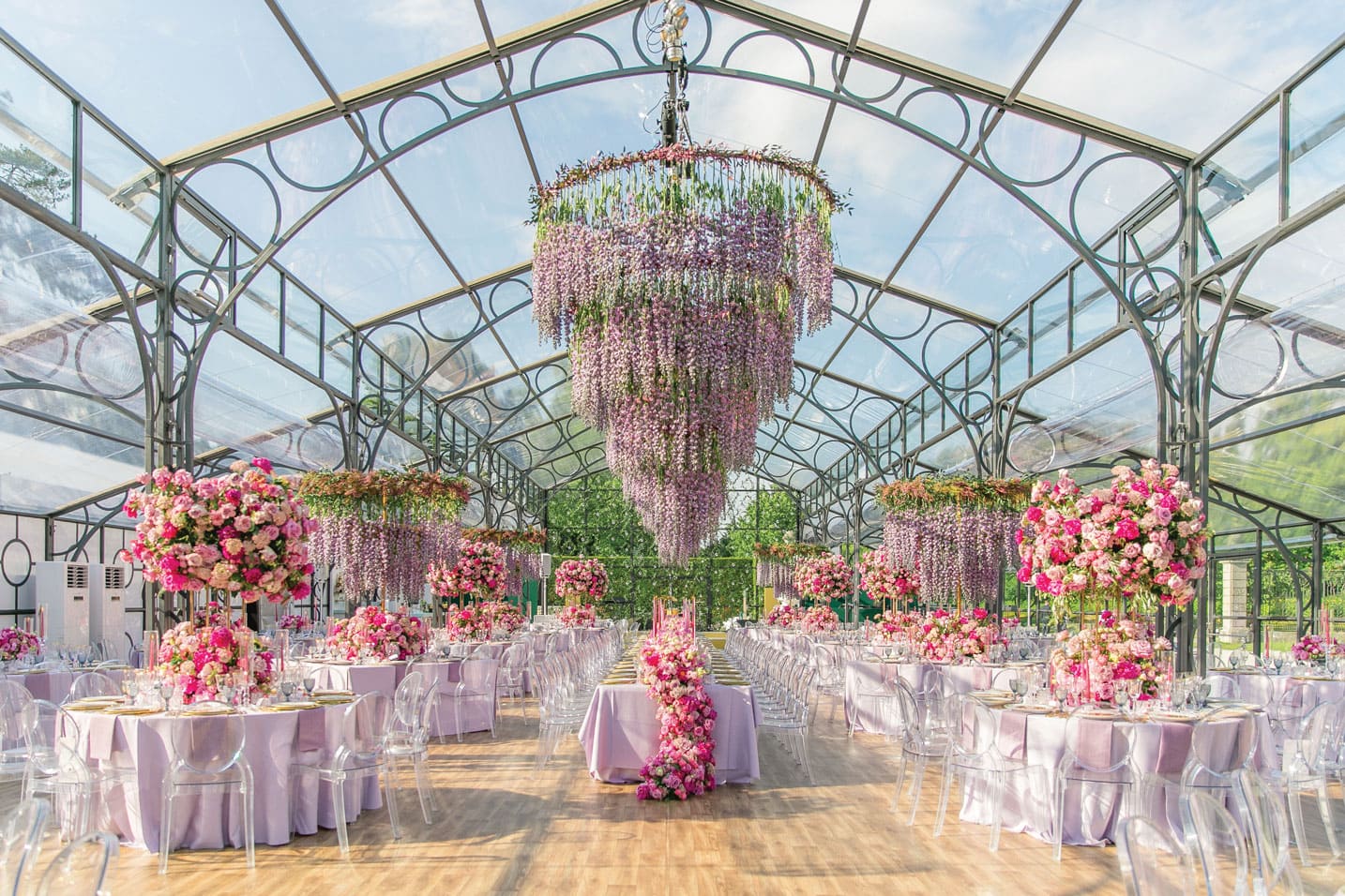 Elegant wedding reception setup with floral centerpieces and hanging flowers in a glass greenhouse.
