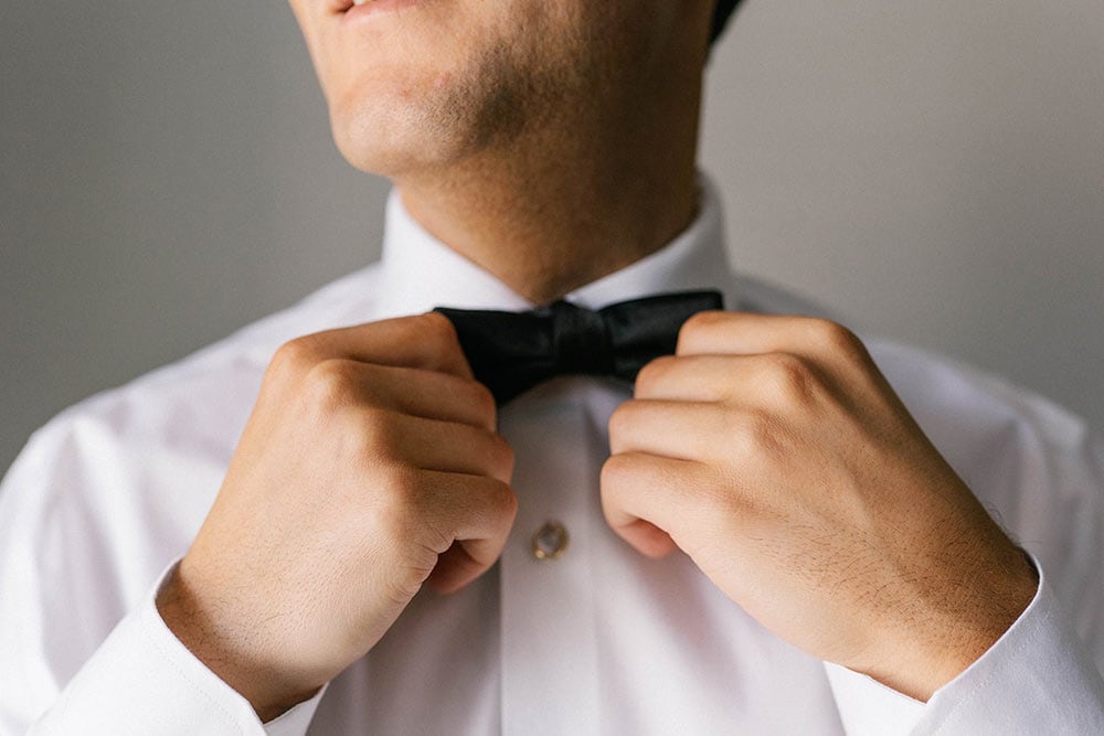 A man adjusting his black bow tie, dressed in a white shirt for a wedding or formal event.