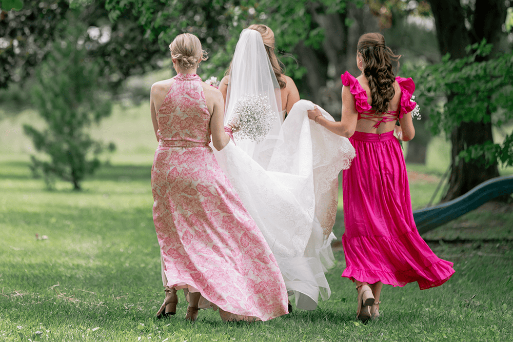 A woman in a wedding dress walking with two young girls in pink dresses outdoors in a park setting.