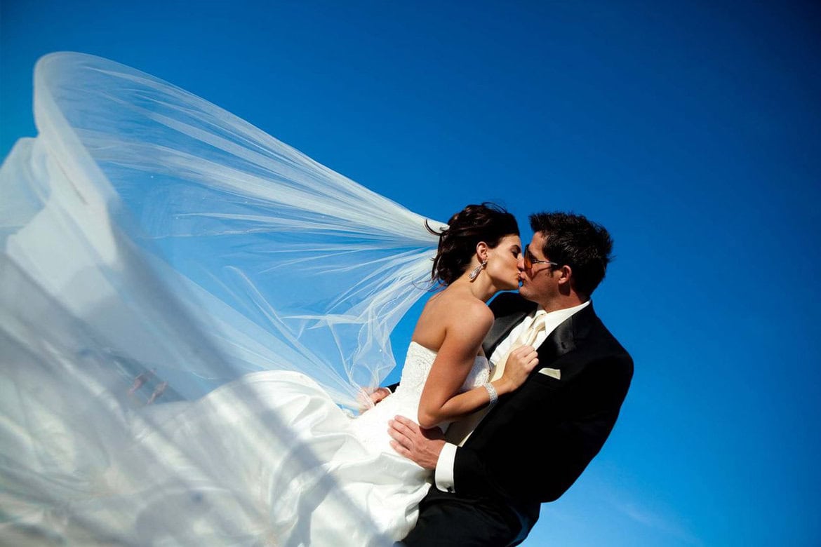 Bride and groom sharing a kiss with flowing wedding veil against blue sky.