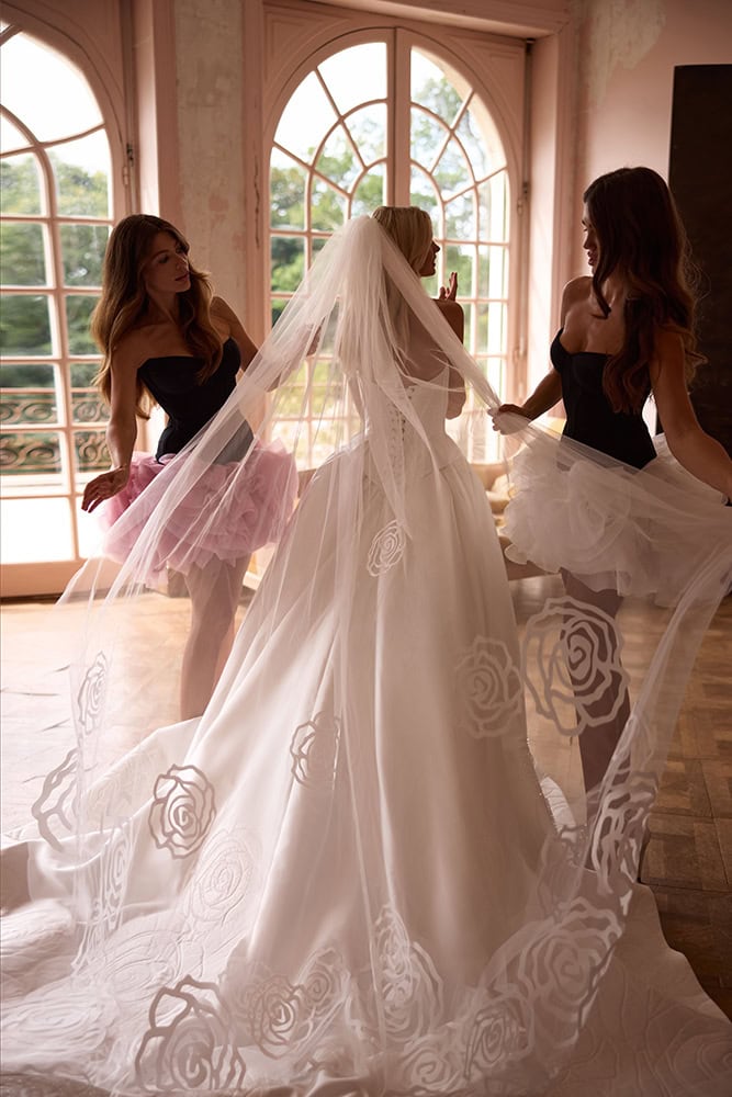 Bride in wedding gown with two bridesmaids holding her veil indoors.