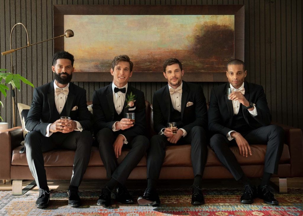 Elegant men in tuxedos sitting on a brown leather sofa at a formal event, holding drinks.