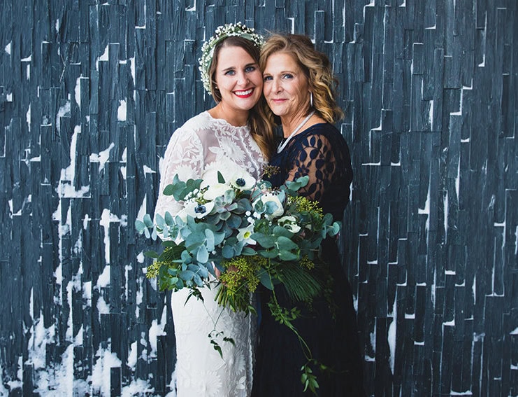 A bride and her mother celebrating wedding day with bouquet against modern dark wall.