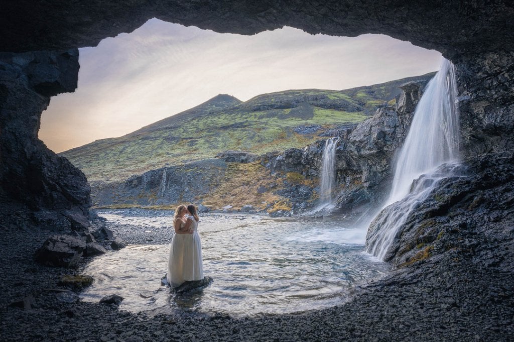 A serene bride standing in a waterfall cave with lush green mountains in the background.