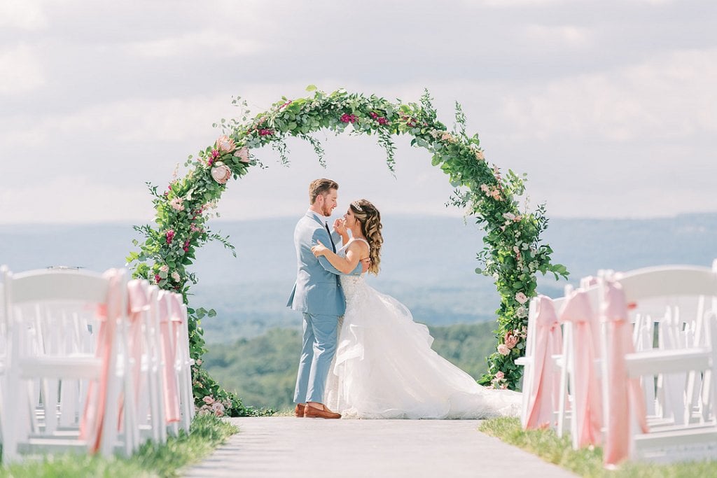 A couple exchanging vows under a floral arch at an outdoor wedding ceremony.