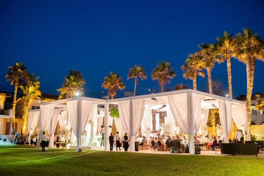 Elegant outdoor wedding reception under a white tent with palm trees at dusk, luxurious event setup.