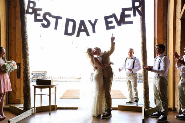 A wedding couple sharing a kiss under "Best Day Ever" banner with friends and family celebrating.