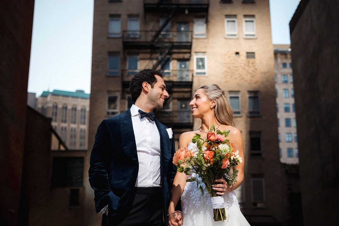 Bride and groom holding hands in urban setting with buildings in background.