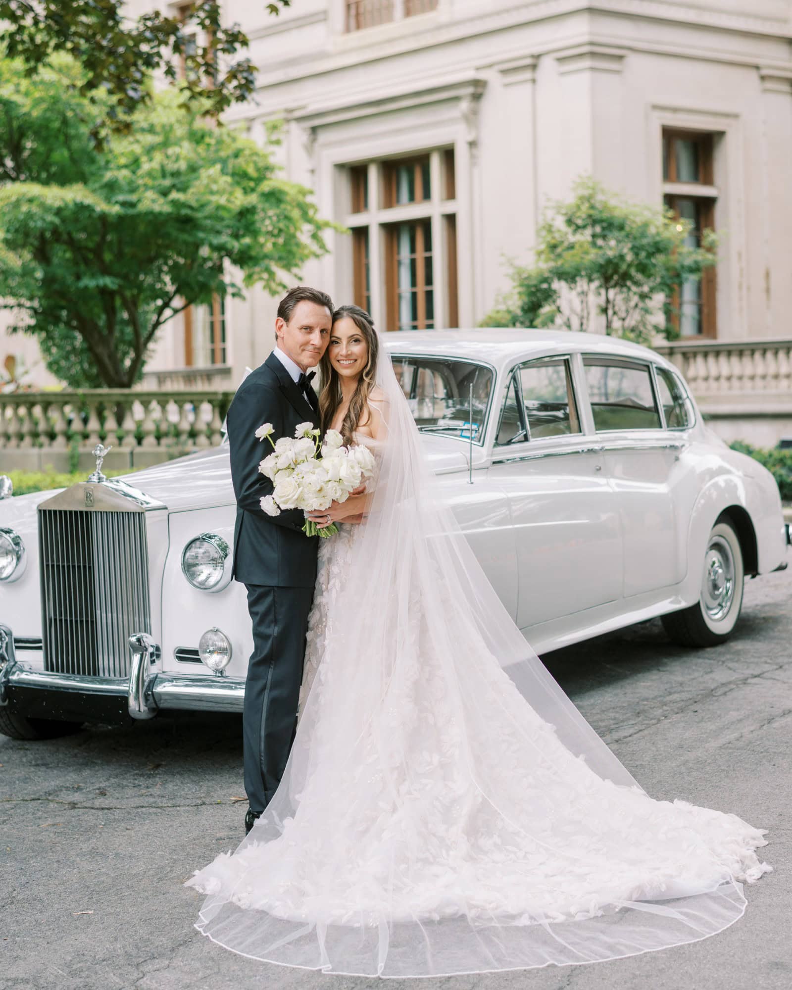 A happy bride and groom with a vintage wedding car outside a historic building.