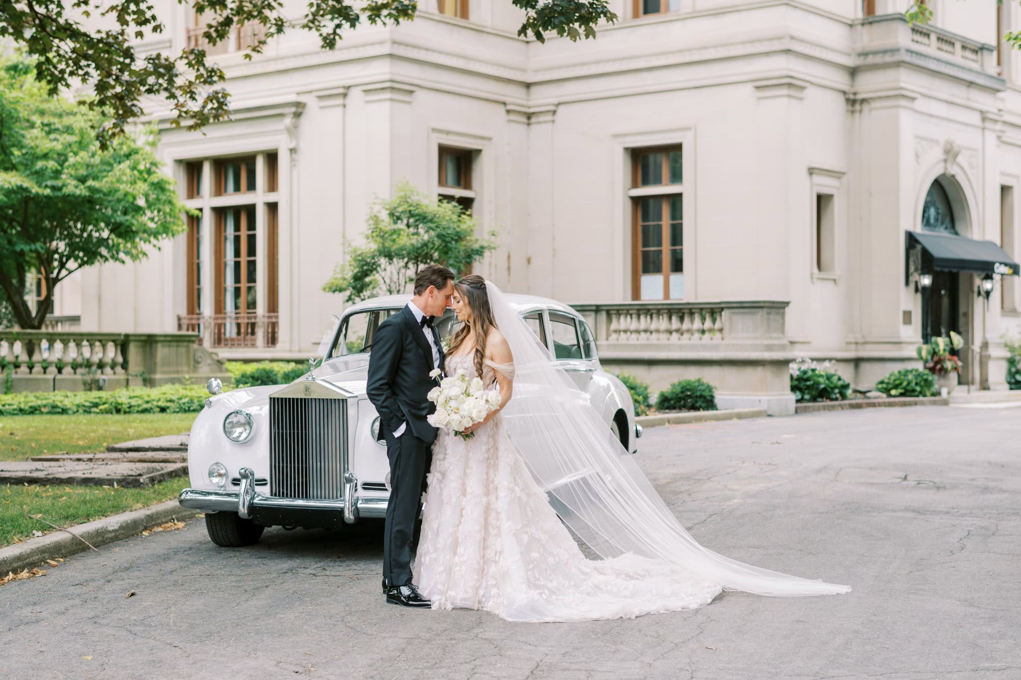 A bride and groom kissing next to a vintage wedding car outside a historic building.