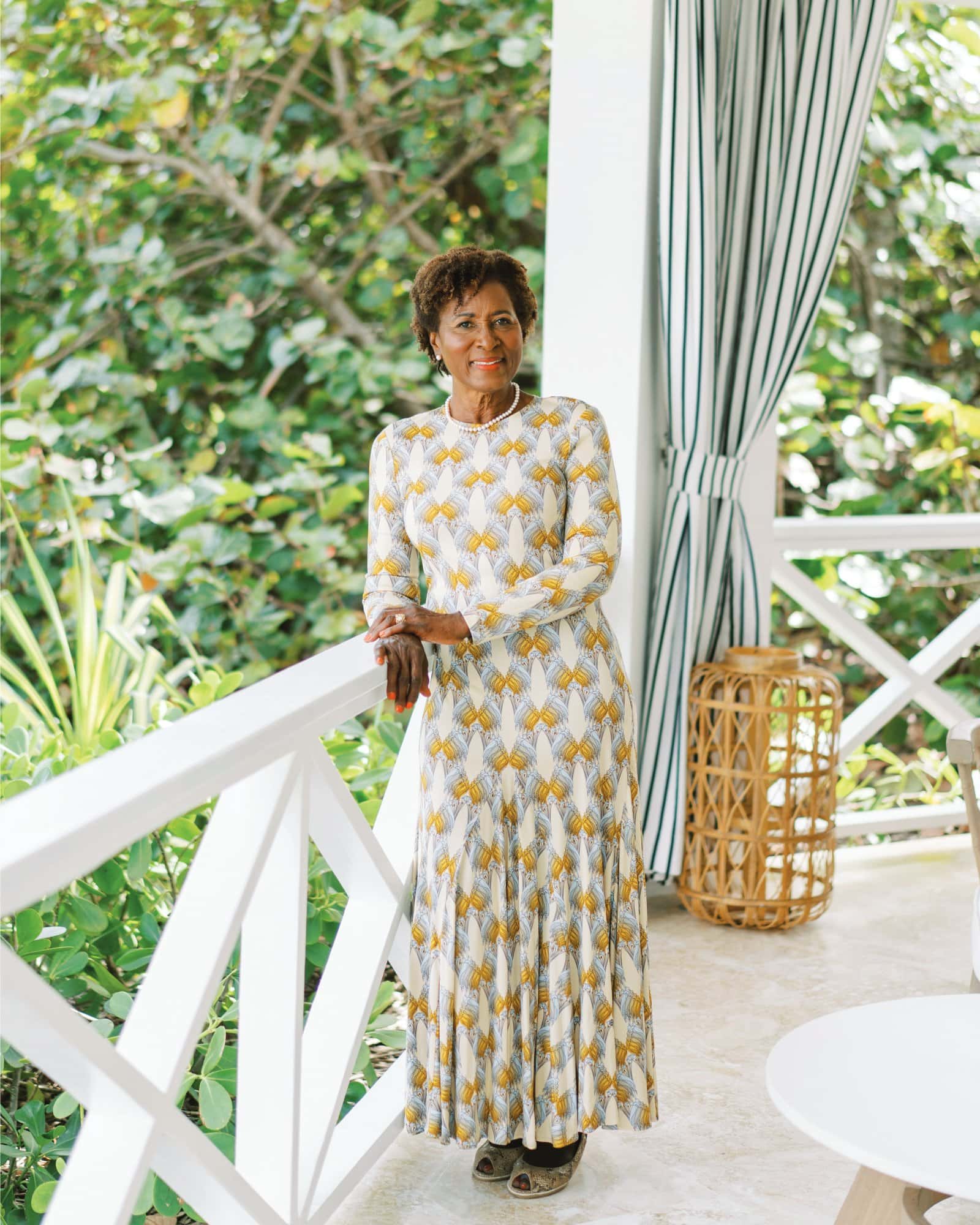 Elegant woman in a floral dress standing on a porch with greenery in the background.