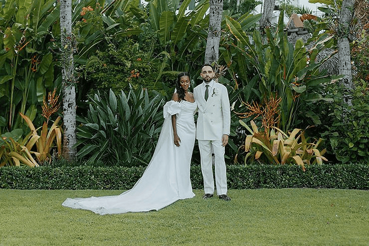 Bride and groom in wedding attire standing outdoors in lush garden setting.