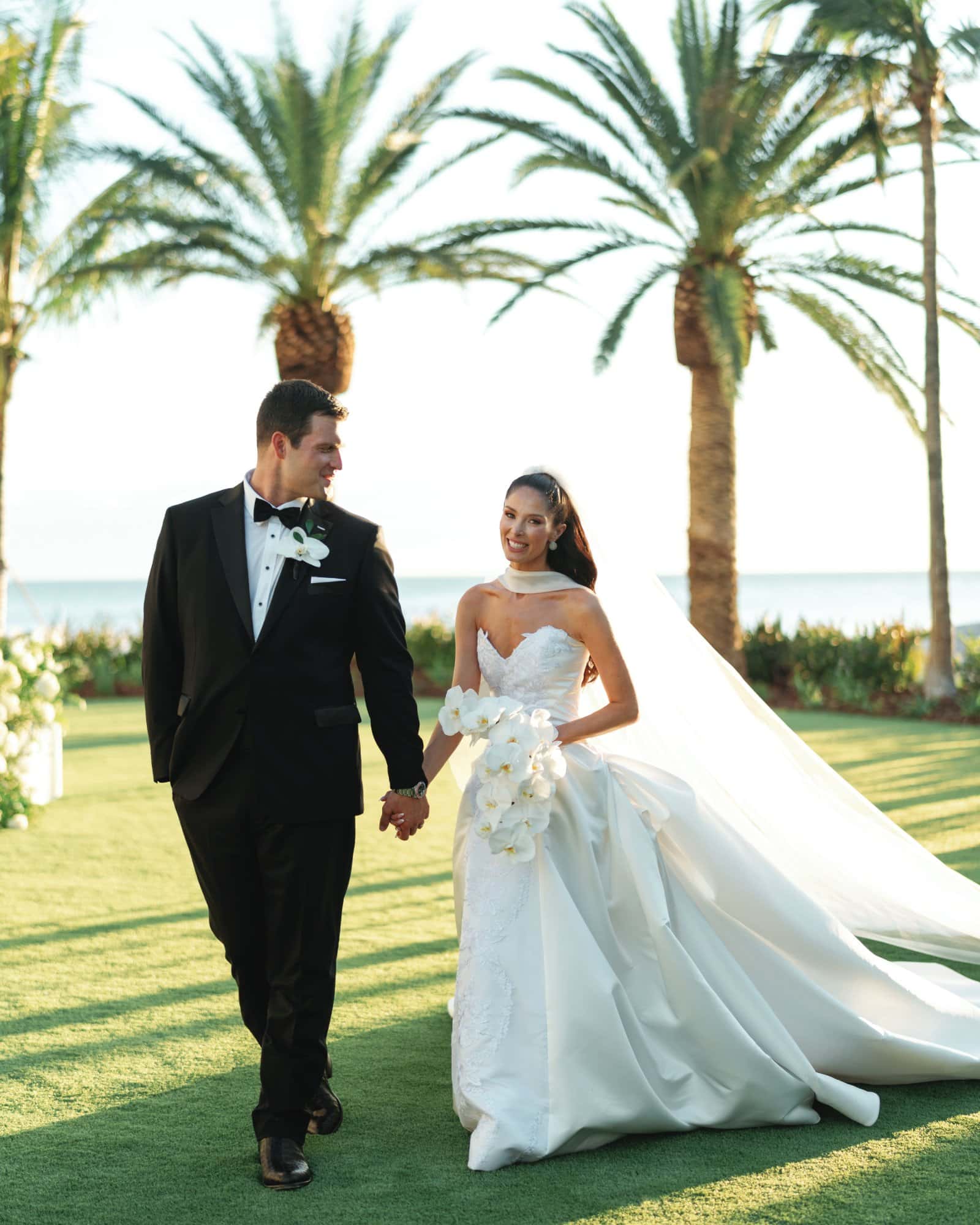 Bride and groom holding hands on a sunny tropical lawn with palm trees.