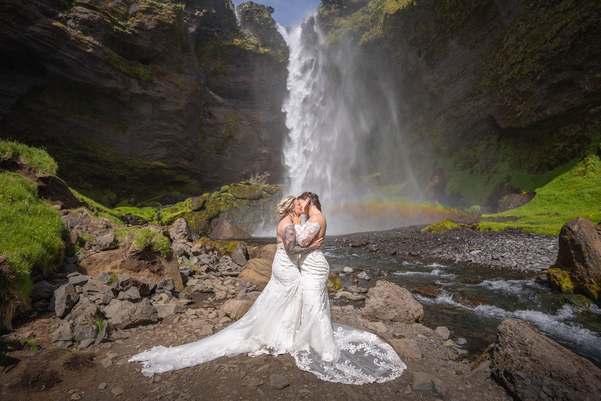 Bride and groom kiss in front of a waterfall at a scenic outdoor wedding.