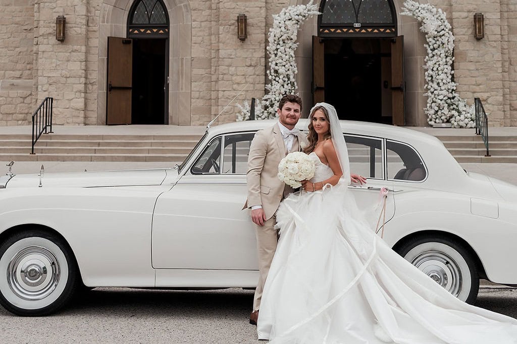 Bride and groom pose with vintage white car outside church after wedding ceremony.