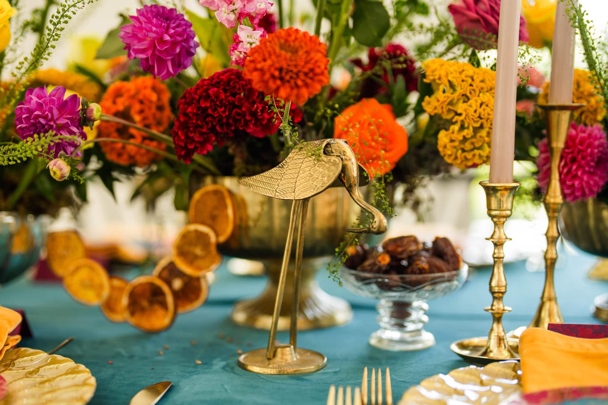 Colorful floral wedding centerpiece with gold accents and candles on a teal tablecloth.