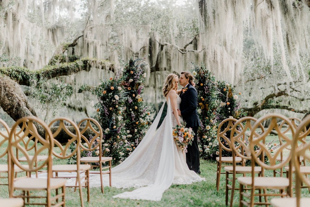 A bride and groom exchanging vows outdoors under Spanish moss-draped trees with floral arch.