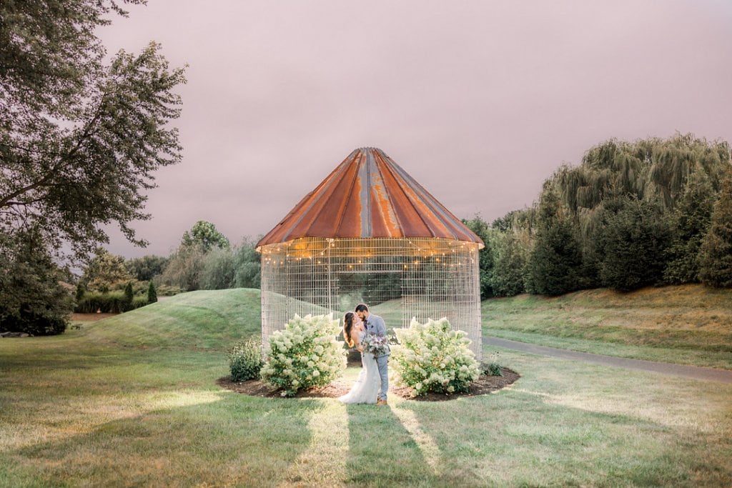 A couple sharing a kiss under a glass and rusted metal pavilion at sunset.