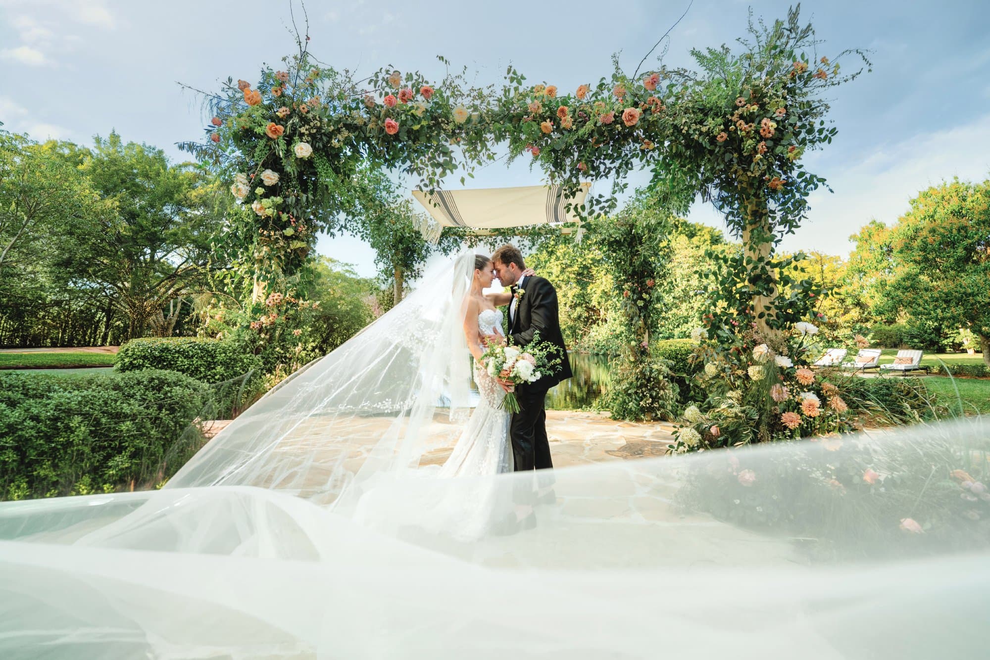 Bride and groom share a kiss under a floral arch at an outdoor wedding ceremony.