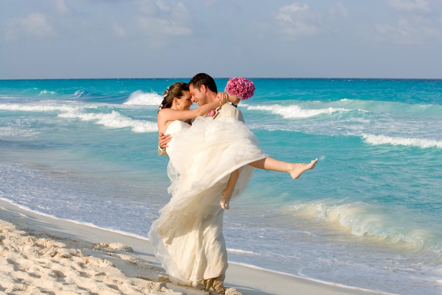 Bride and groom embrace on the beach with the ocean in the background.