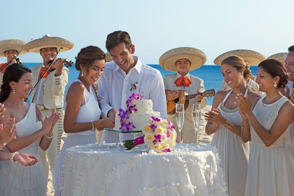 Bride and groom cutting wedding cake on beach with friends and mariachi band.