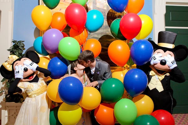 Bride and groom kissing among colorful balloons with Mickey and Minnie Mouse characters.