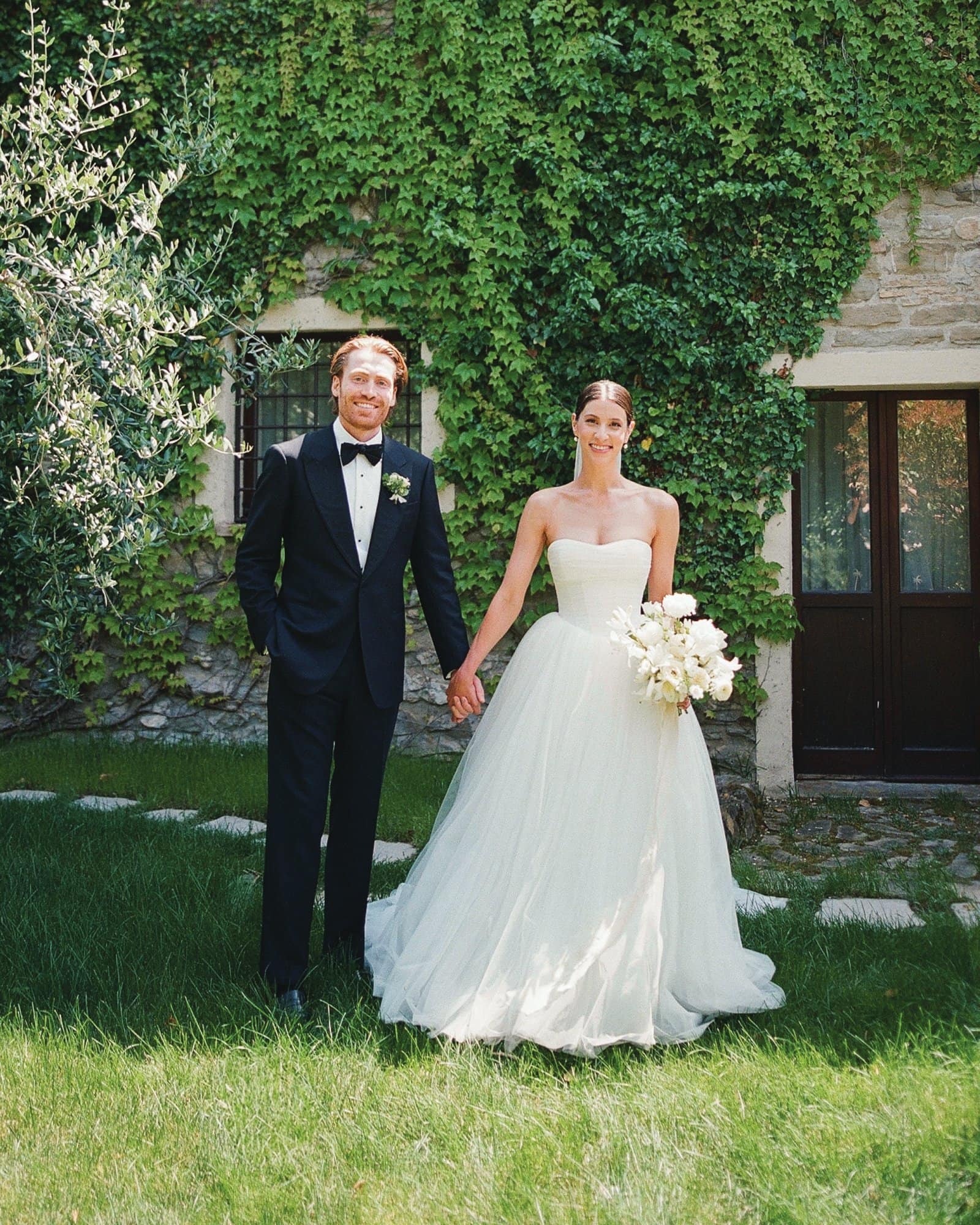 Bride and groom holding hands outside in wedding attire with greenery background.