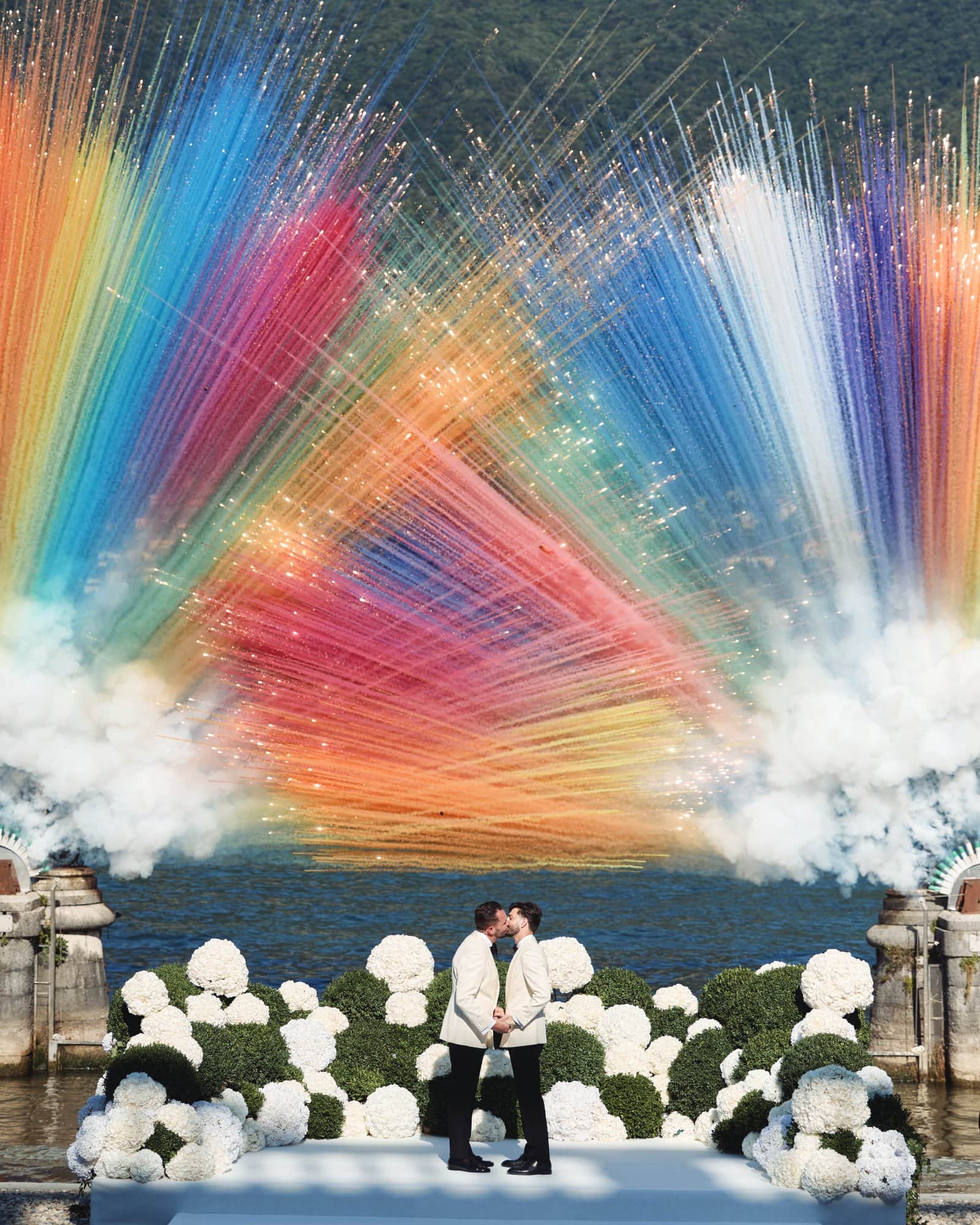 Couple kissing at a colorful fireworks display over a lake during a wedding celebration.