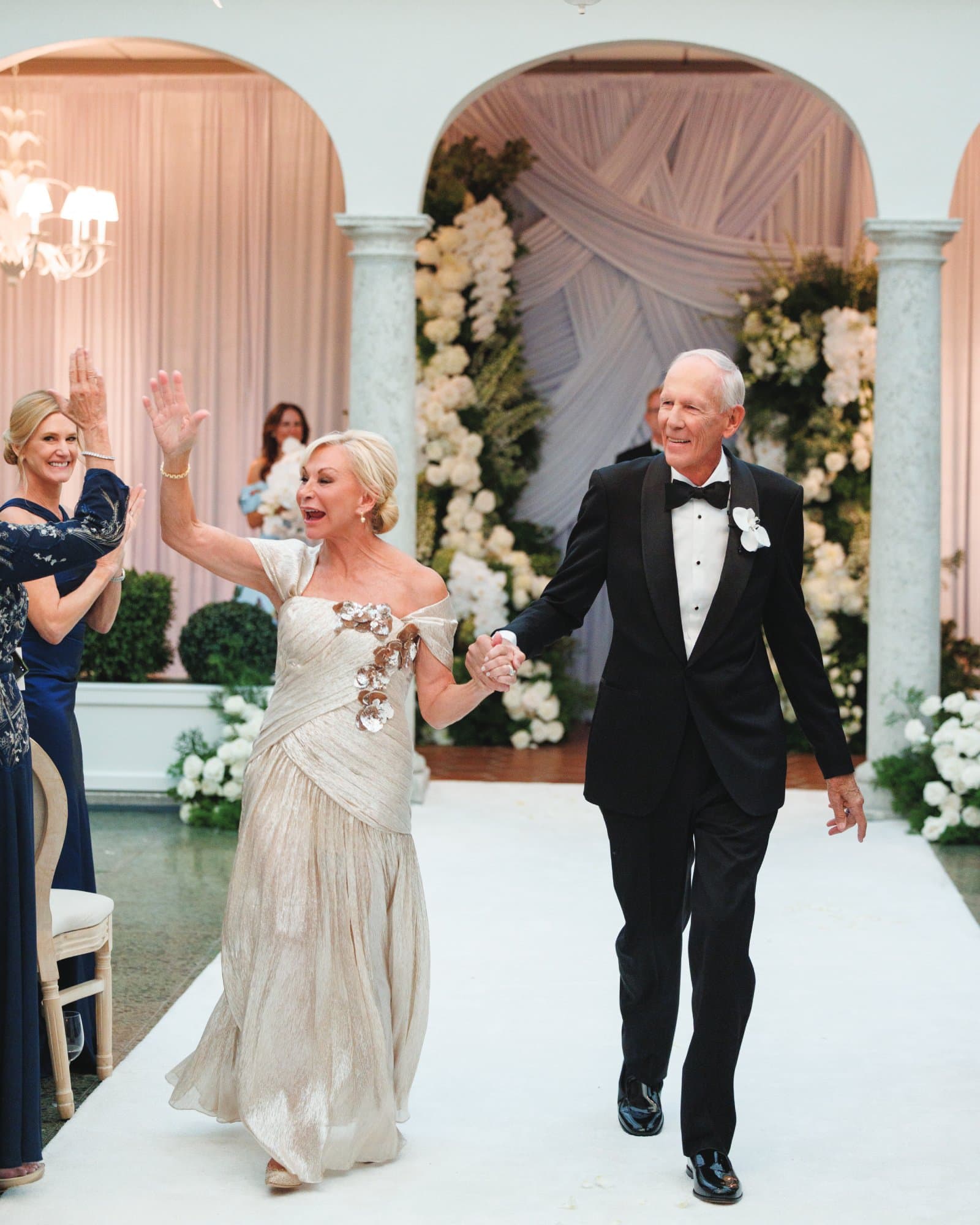 Elderly couple dancing at wedding reception with floral decor and guests in background.
