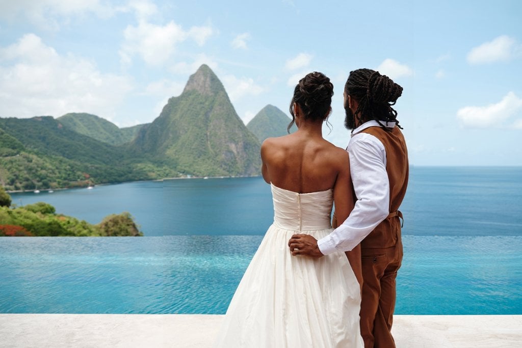 Couple in wedding attire enjoying scenic ocean view with mountains in the background.