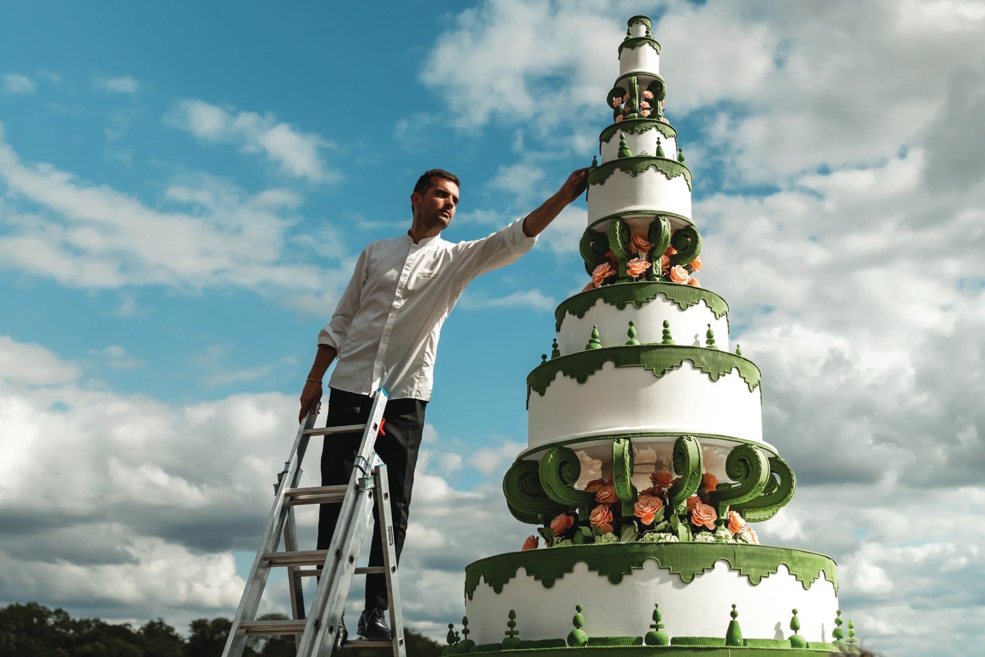 Man decorating a large wedding cake outdoors with a ladder and blue sky.